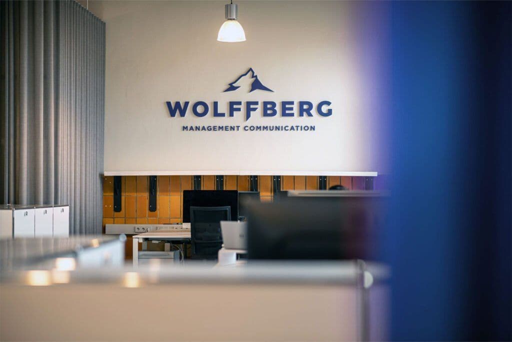 Blick in ein Großraumbüro bei WOLFFBERG. Im Anschnitt rechts ein blauer Vorhang, hinten sieht man Monitore auf Tischen stehen. An der Wand hinten steht der Schriftzug WOLFFBERG Manamgement Communication.