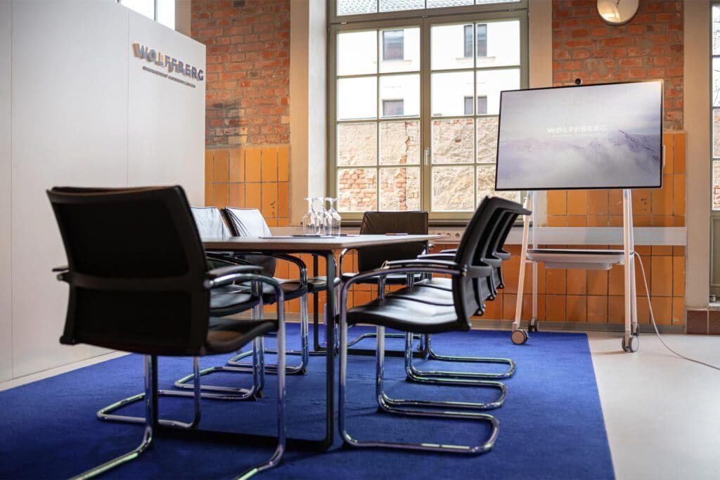Blick in einen Konferenzraum bei WOLFFBERG Management Communications. In der Mitte steht ein ovaler Tisch mit mehreren Stühlen auf einem blauen Teppich. Vor den großen Fenster im Hintergrund steht ein großer Bildschirm.